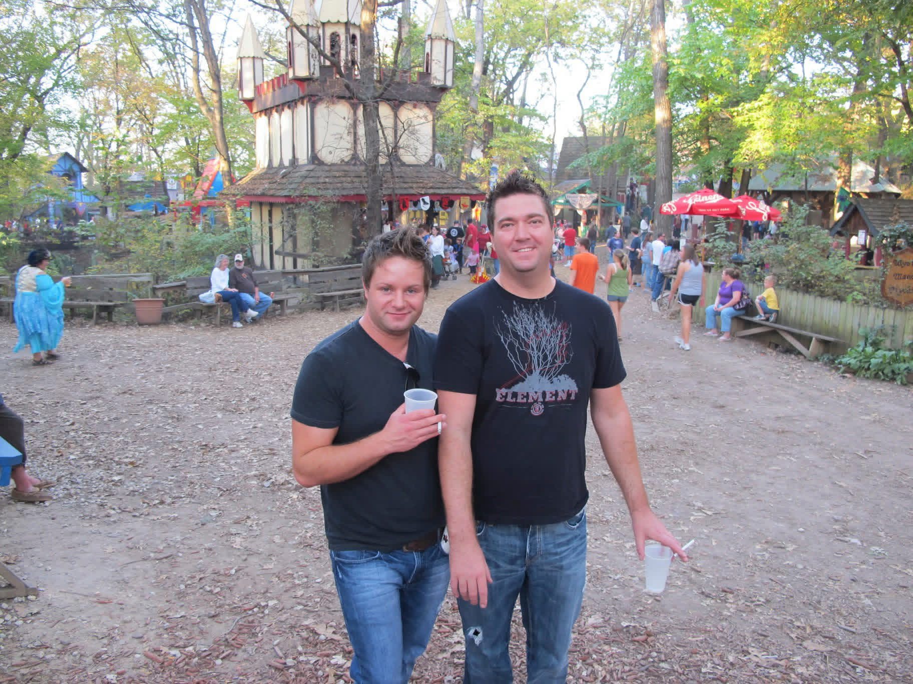 Shane (right) with his friend Ian at a Renaissance festival, a wooden castle behind them.