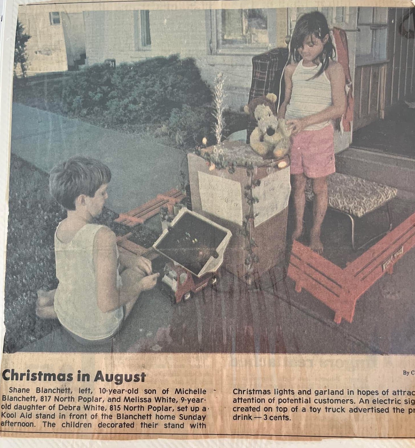 Newspaper clipping headlined "Christmas in August": a ten-year-old Shane and a neighbor run a Kool-Aid stand decorated with a little Christmas tree and lights outside his family's home.