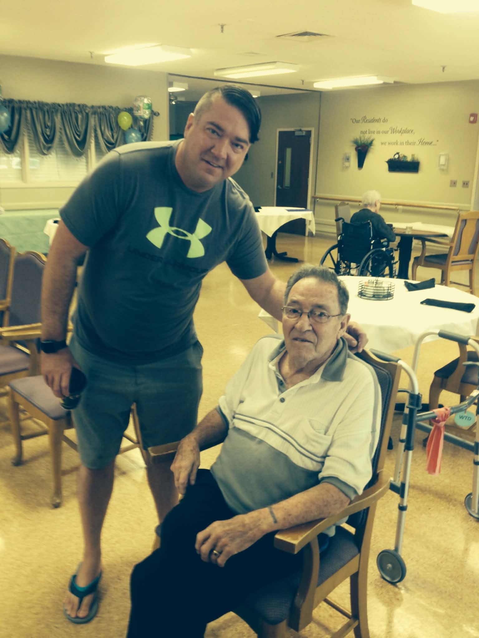 Shane standing beside his grandpa Leo, who is seated, during a visit to Leo's care home.